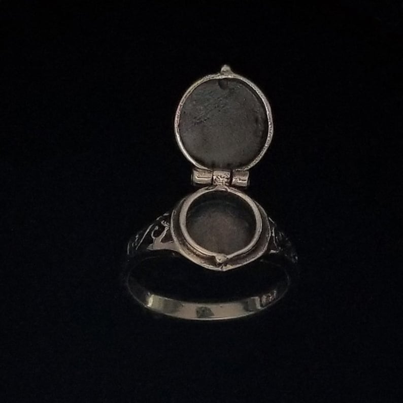 Op de afbeelding: Een zilveren ring met een scharnierend, rond medaillon dat opengaat om een lege ruimte binnenin te onthullen. De ring heeft een ingewikkeld ontwerp rond het medaillon.