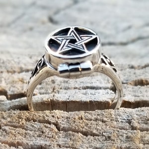 Op de afbeelding: Een zilveren ring met een scharnierend, rond medaillon met een pentagram-ontwerp. De ring is versierd en heeft een gedetailleerd ontwerp.