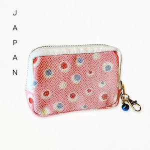 May include: A rectangular zippered pouch featuring a textured pink fabric with a pattern of small red, white, and blue circles. The pouch has a white trim and a gold-toned metal lobster clasp attached to a small blue bell charm on the side.