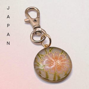 May include: A gold-coloured keychain with a round pendant featuring a floral design. The pendant has a clear background with a pink flower and gold and green accents. The word "JAPAN" is printed vertically on the left.