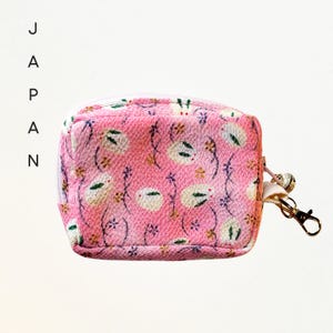 May include: A small pink zippered pouch featuring a repeating pattern of white rabbits and small floral sprigs. The pouch includes a gold-toned metal lobster clasp and a small bell attached to the side zipper pull, with the word JAPAN printed vertically.