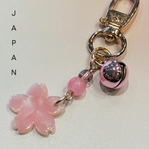 May include: A gold-coloured keyring featuring a pink bell, pink beads, and a pink flower charm. The word "JAPAN" is printed vertically on the left. The keyring is designed with a spring clasp.
