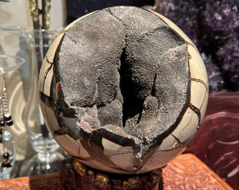 May include: A geode sphere with a natural crack revealing the interior crystals. The sphere is displayed on a dark red wooden stand with three legs.