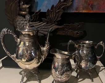 May include: A silver tea set with a teapot, creamer, and sugar bowl. The set has an ornate design and is on a white surface.