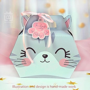 Op de afbeelding: Een lichtblauwe katvormige geschenkdoos met een roze lint en label. De doos heeft een kattengezicht met roze wangen en zwarte snorharen. Op het label staat "Thank you for coming! 5th Birthday Amelia."