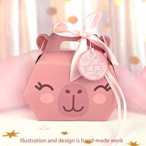 Puede incluir: Una caja de regalo rosa hexagonal con un diseño de cara de llama de dibujos animados. La caja tiene un asa y está atada con una cinta y una etiqueta rosas. La etiqueta dice "5th Birthday Amelia Thank you for celebrating!" El texto "Illustration and design is hand-made work" está en la parte inferior.
