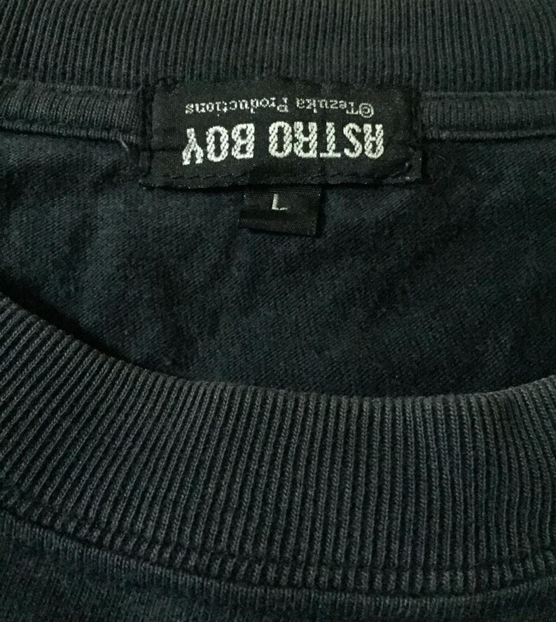 May include: A black Astro Boy t-shirt tag with white text that reads "Astro Boy" and "&copy;Tezuka Productions". The tag also has a white size label that reads "L".