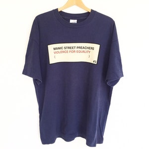 May include: A navy blue t-shirt with a white rectangle featuring the text "Manic Street Preachers (Violence for Equality) #3" in black and red lettering.
