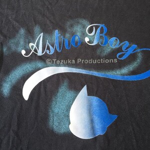 May include: A black t-shirt with a white and blue spray-painted design that says "Astro Boy" with the copyright symbol and "Tezuka Productions" underneath. A blue and white silhouette of Astro Boy's head is in the lower right corner.