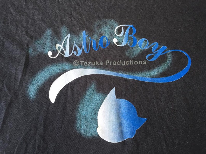 May include: A black t-shirt with a white and blue spray-painted design that says "Astro Boy" with the copyright symbol and "Tezuka Productions" underneath. A blue and white silhouette of Astro Boy's head is in the lower right corner.