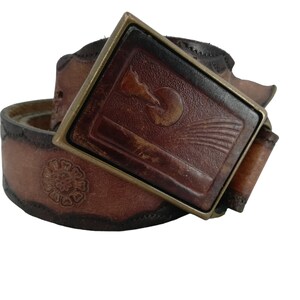 May include: A brown leather belt with a decorative buckle featuring a sun and a bird design. The belt has a stitched edge and a floral design.