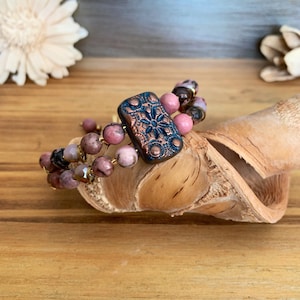 Könnte beinhalten: Ein mehrreihiges Armband mit rosa und braunen Perlen und einem rechteckigen Anhänger in Kupferton mit floralem Design.