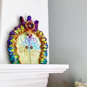 Puede incluir: Una puerta de hadas caprichosa con un marco morado, azul y verde adornado con alambre dorado y cristales. La puerta está decorada con una guirnalda de cuentas y tiene un fondo blanco con un diseño azul y blanco.