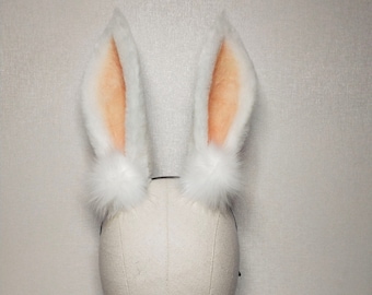 May include: White bunny ears with a fluffy texture and peach-colored inner ears. The ears are attached to a black headband and have fluffy white pom-poms at the base. The ears are standing upright.