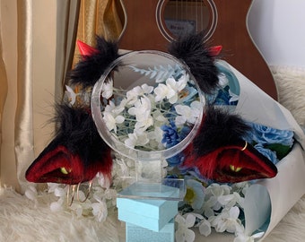 May include: A pair of red devil horns with black fur and a red velvet base. The horns are on a clear plastic stand with a white and blue floral background.