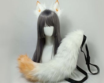 May include: A white and orange fox tail with a black strap and buckle, paired with matching white fox ears with orange tips. The ears are attached to a gray wig on a mannequin head. The tail is fluffy and detailed.