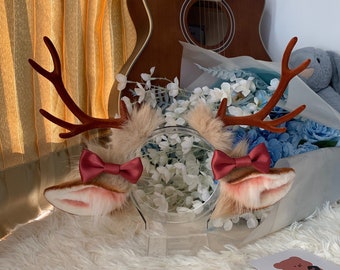 May include: A pair of deer antlers and ears headband. The antlers are a dark brown color, and the ears are a light brown with pink accents. A burgundy bow tie is attached to each ear. The headband is displayed on a clear stand.