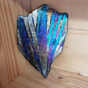 May include: An abstract art piece with a jagged edge, displaying a spectrum of colours including blues, purples, and silver. The textured surface gives a layered effect. The artwork is set against a wooden backdrop, enhancing its visual appeal.