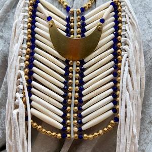May include: A decorative chest piece featuring rows of cream-colored cylindrical beads, vibrant blue glass beads, and metallic gold-toned spherical beads. A large, smooth, crescent-shaped brass pendant sits at the top, framed by long white leather fringe.