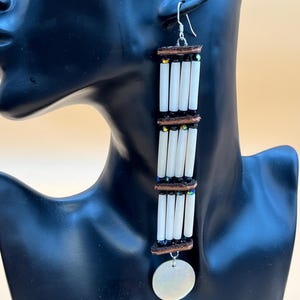 May include: Handmade dangle earrings featuring white cylindrical beads, dark brown accents, and iridescent beads. A circular, pearlescent charm hangs at the bottom. The earrings are suspended from silver-colored hooks. The earrings are displayed on a black mannequin.