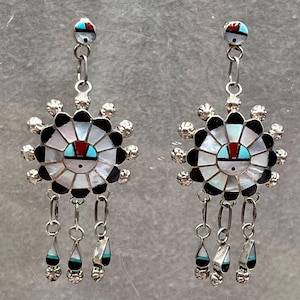 May include: A pair of silver earrings with a sunface design. The earrings feature a central sunface with a turquoise, red, and black design, surrounded by white and black segments. Dangling from the main design are three teardrop-shaped charms with turquoise and black accents.