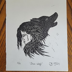 Peut inclure: Une estampe en noir et blanc représentant une femme avec une tête de loup. Le visage de la femme est visible et elle regarde vers le bas. La tête du loup est tournée sur le côté et sa gueule est ouverte. L'estampe est signée 'She-Wolf' et 'DB 09/24'.