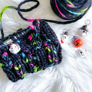 Pode incluir: Uma bola de lã preta, verde e rosa com um chapéu de malha preto e branco com detalhes de lã coloridos. O chapéu é decorado com pequenos pingentes temáticos de Halloween.