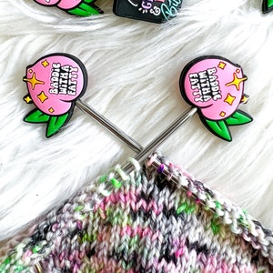 May include: A close-up of a set of colorful peach-shaped charms with various phrases, including "Beauty & Grace & Resting Bitch Face" and "Baddie With a Fatty". The charms are on a white fuzzy surface with knitting needles and a multi-colored knitted fabric.