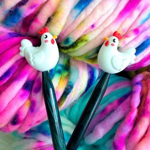May include: Two black crochet hooks featuring white chicken-shaped toppers with red combs and yellow beaks. The chicken figures have small black eyes and pink cheeks. The hooks are positioned against a background of multi-coloured tie-dye yarn.
