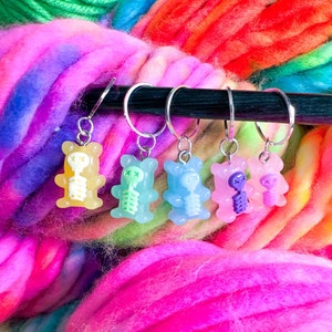 May include: Six colorful gummy bear stitch markers with skeleton designs. The markers are attached to a silver metal ring and are hanging from a wooden dowel. The markers are in shades of yellow, green, blue, purple, and pink.