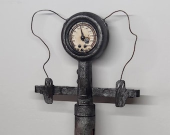 May include: A steampunk-style metal syringe with a gauge attached to the top. The syringe is silver with a black and red design. The gauge is black with a white face and black numbers. The syringe is mounted on a black metal stand.