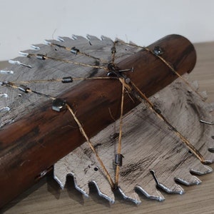 Puede incluir: Una hoja de sierra circular con dientes, unida a un mango de madera con cuerda y sujetadores metálicos. La hoja está pintada de plata y el mango es marrón.