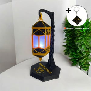 Può includere: Una lanterna nera e dorata con interno blu brillante, sostenuta da un supporto nero attorcigliato. La base del supporto riporta la scritta "EXPEDITION" e il numero "33" in lettere dorate. È incluso un portachiavi bonus.