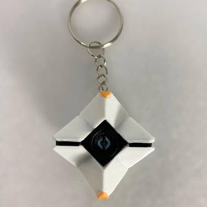 Op de afbeelding: Witte en zwarte geometrische sleutelhanger met een zilveren sleutelring. De sleutelhanger heeft een ruitvorm met een zwart midden en oranje accenten. Het midden heeft een blauw ontwerp.