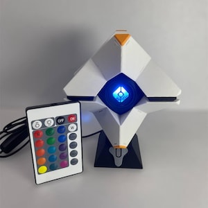 Op de afbeelding: Een witte en zwarte geometrische lamp met oranje accenten en een blauw licht in het midden. Een afstandsbediening met kleuropties staat links. De lamp staat op een zwarte basis.