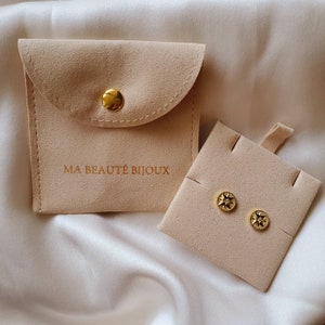 Puede incluir: Dos pendientes de estrella dorados con una pequeña piedra transparente en el centro, presentados en una caja de joyería de terciopelo beige con el texto "MA BEAUTÉ BIJOUX".