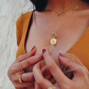 May include: A gold chain necklace with a large coin pendant featuring a face and a toggle clasp. The necklace is layered over a thinner gold chain with a small bar pendant. The person is wearing a mustard yellow top and has their hands in the frame.
