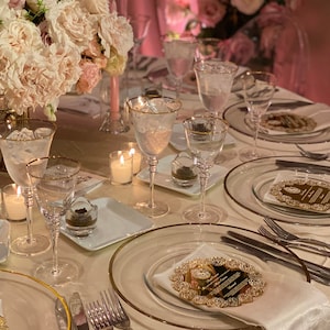 May include: A table setting with gold-rimmed glassware, white plates, and gold-rimmed place cards. The table is decorated with pink flowers and candles.