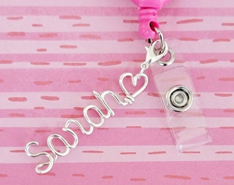May include: A silver name badge holder with the name "Sarah" spelled out in wire with a heart at the end. The holder is attached to a pink heart-shaped retractable badge reel.