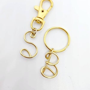 May include: Gold-colored metal keychains with the letters "S" and "B". One keychain has a lobster clasp, and the other has a keyring. The letters are in a cursive font.