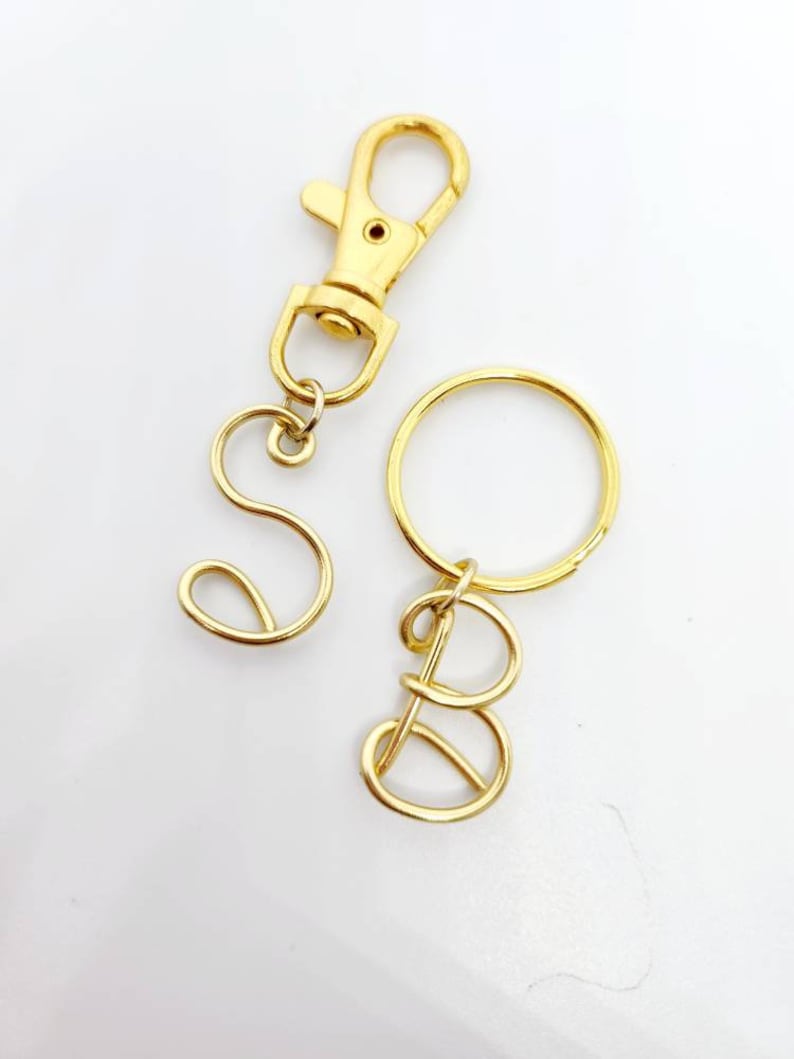 May include: Gold-colored metal keychains with the letters "S" and "B". One keychain has a lobster clasp, and the other has a keyring. The letters are in a cursive font.