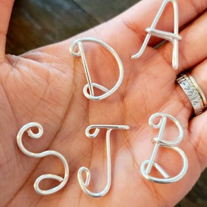 May include: Close-up of silver wire letters spelling out the initials S, J, B, D, and A. The letters are handmade and have a delicate, cursive style. The letters are displayed on a person's open palm.