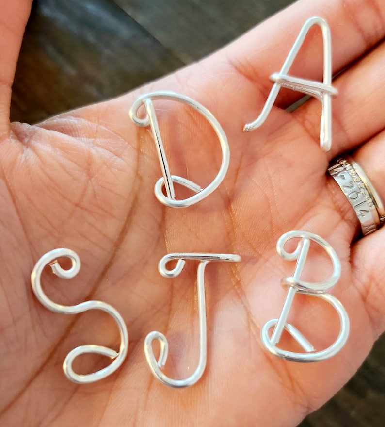 May include: Close-up of silver wire letters spelling out the initials S, J, B, D, and A. The letters are handmade and have a delicate, cursive style. The letters are displayed on a person's open palm.