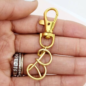 May include: A gold-colored swivel clasp with a lobster claw closure and two connected key rings. The metal hardware is shiny and appears to be new. The clasp is held in a hand with multiple rings on the fingers.