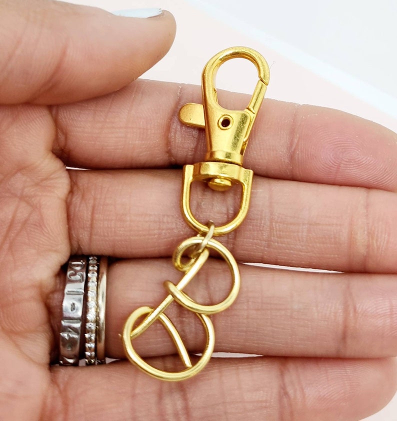 May include: A gold-colored swivel clasp with a lobster claw closure and two connected key rings. The metal hardware is shiny and appears to be new. The clasp is held in a hand with multiple rings on the fingers.