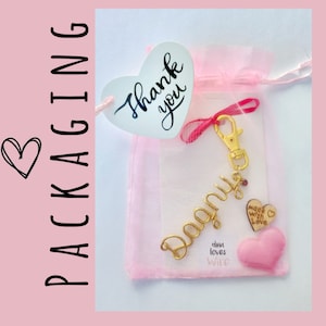 May include: A gold-colored keychain with the name "Donna" spelled out in cursive. The keychain is packaged in a pink organza bag with a heart-shaped tag that says "Thank you". The bag also includes a small pink heart with the text "Made with Love" and a smaller pink heart with the text "Dina loves Wire".