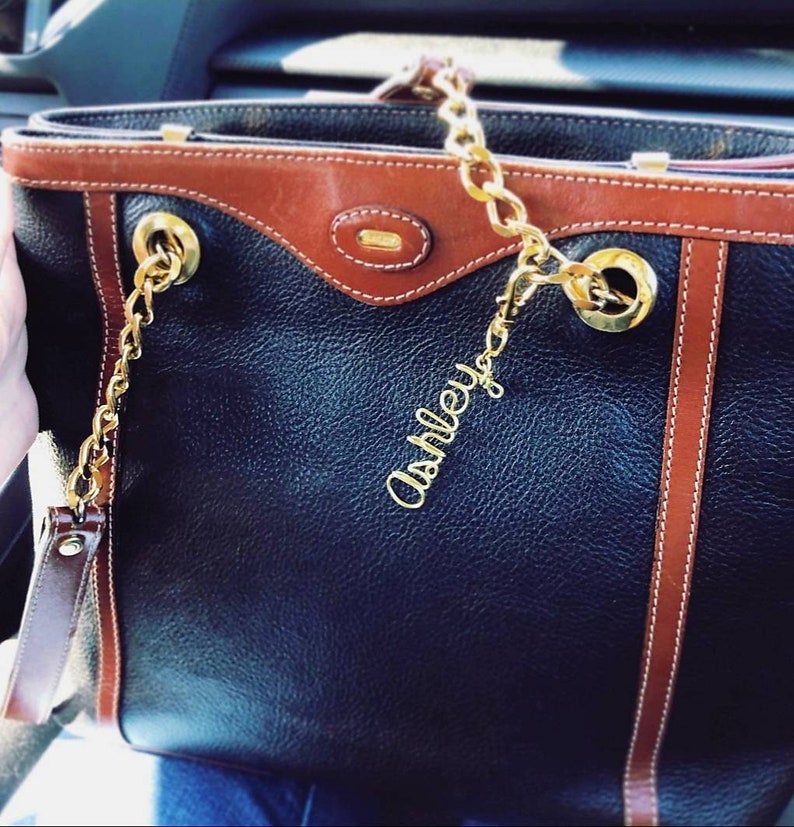May include: A black leather tote bag with brown leather trim and a gold chain strap. The bag has a gold name tag that says "Ashley".