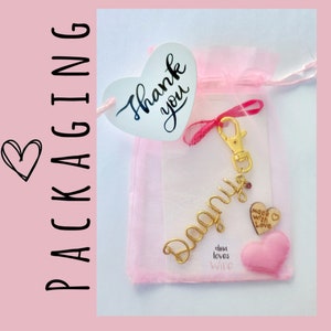 May include: A gold-colored keychain with the name "Donna" spelled out in cursive. The keychain is packaged in a pink organza bag with a heart-shaped tag that says "Thank you". The bag also includes a small pink heart with the text "Made with Love" and a smaller pink heart with the text "Dina loves Wire".