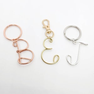 May include: Three wire letter keychains. The letters are B, C, and J. The letter B is rose gold, the letter C is gold, and the letter J is silver. Each letter is attached to a keyring or clasp.