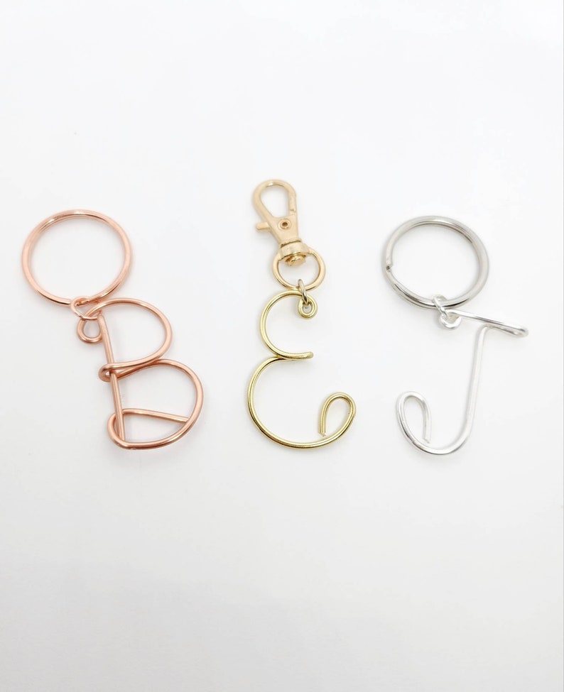 May include: Three wire letter keychains. The letters are B, C, and J. The letter B is rose gold, the letter C is gold, and the letter J is silver. Each letter is attached to a keyring or clasp.